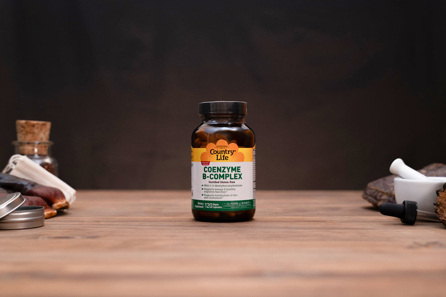 Country Life Coenzyme B-Complex supplement bottle on a wooden tabletop with a dark, moody background and small herbal props on either side.