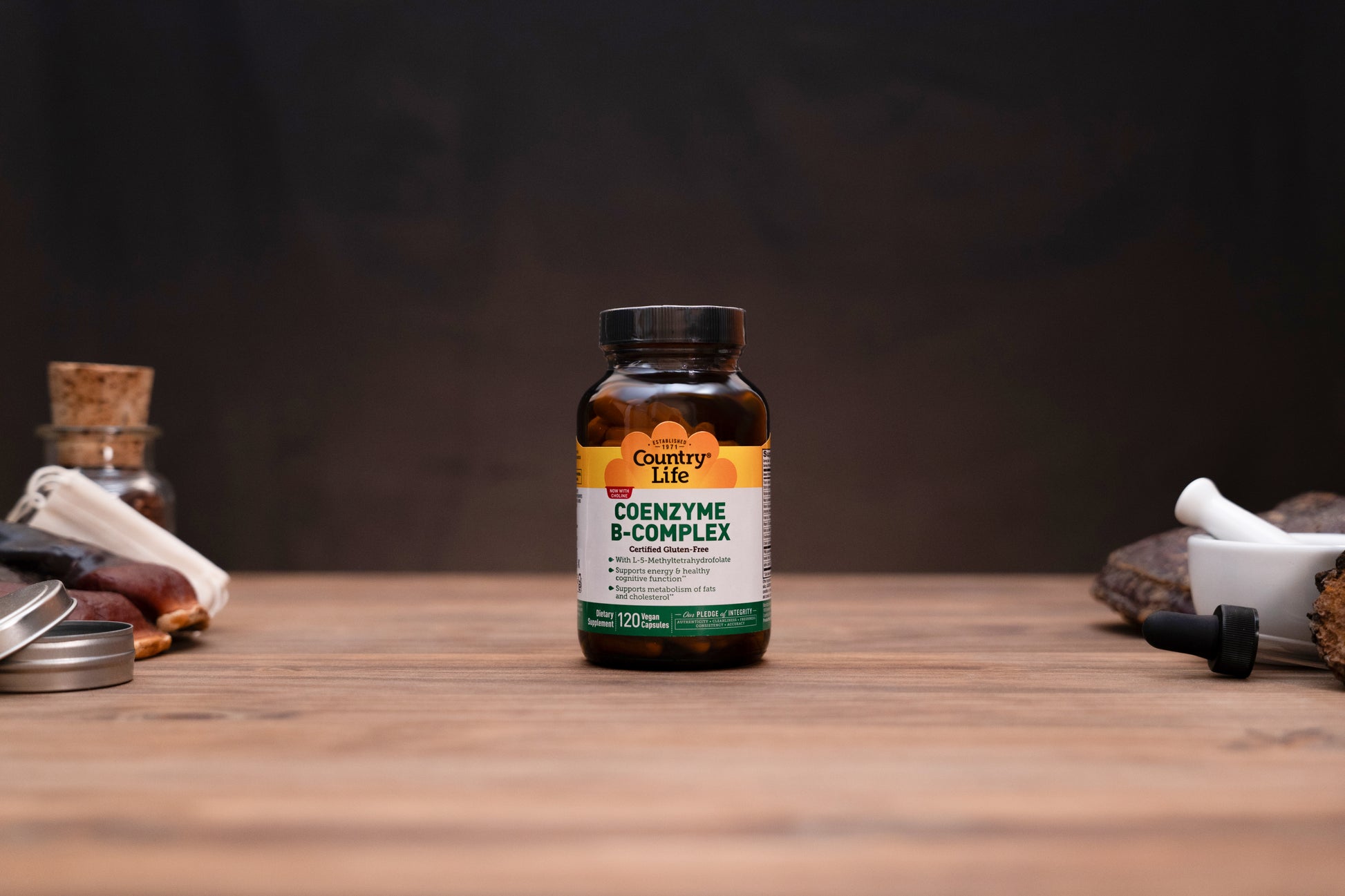 Country Life Coenzyme B-Complex supplement bottle on a wooden tabletop with a dark, moody background and small herbal props on either side.