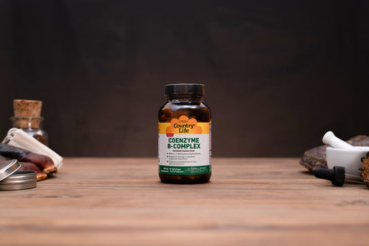 Country Life Coenzyme B-Complex supplement bottle on a wooden tabletop with a dark, moody background and small herbal props on either side.