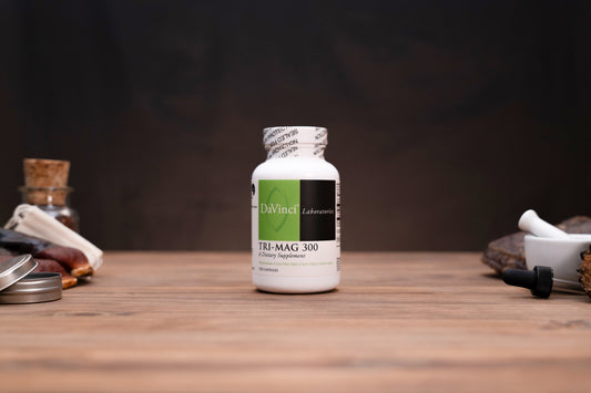 A white bottle of DaVinci Lab Tri-Mag 300 dietary supplement stands centered on a rustic wooden table, surrounded by blurred herbal and laboratory props against a dark background.