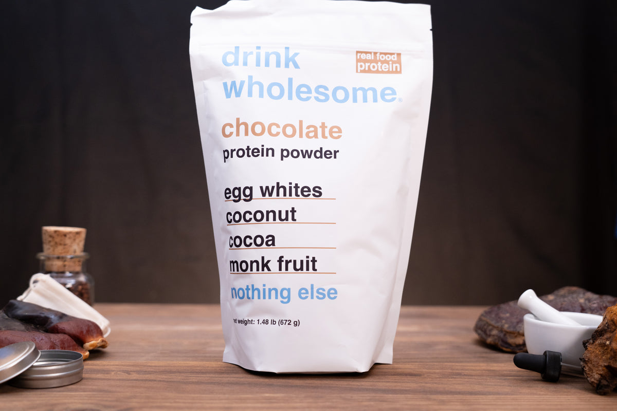 A white stand-up pouch of drink wholesome chocolate protein powder, with ingredients listed on the front, set on a wooden surface with dark background and small food props around it.
