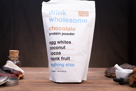 A white stand-up pouch of drink wholesome chocolate protein powder, with ingredients listed on the front, set on a wooden surface with dark background and small food props around it.