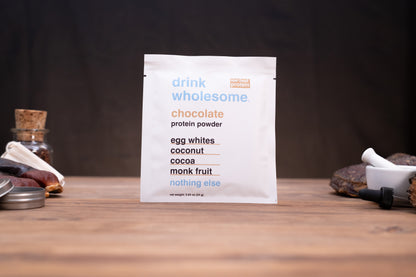 A white packet of chocolate protein powder stands upright on a wooden surface against a dark background. The packet reads 'drink wholesome.