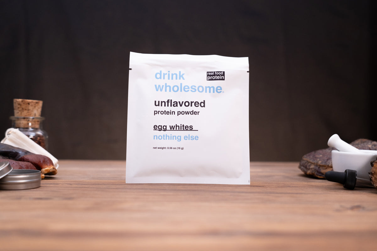 A white packet of drink wholesome unflavored protein powder stands on a wooden surface against a dark background, with small kitchen props visible on both sides.