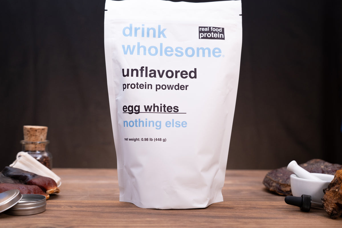 A white stand-up pouch of drink wholesome unflavored protein powder, labeled egg whites, standing on a wooden surface against a dark background with small kitchen props around it.