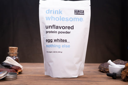 A white stand-up pouch of drink wholesome unflavored protein powder, labeled egg whites, standing on a wooden surface against a dark background with small kitchen props around it.