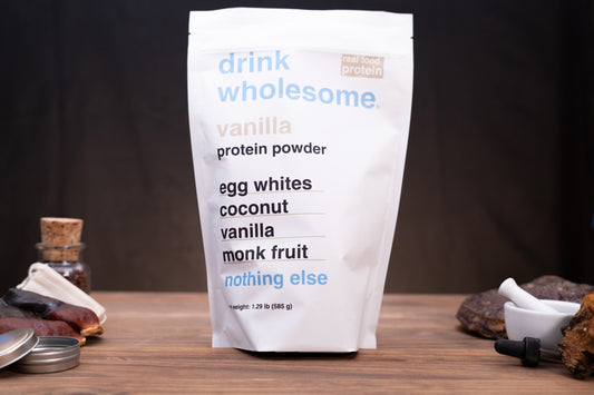 A stand-up pouch of vanilla protein powder from the brand drink wholesome, displayed on a wooden surface with small kitchen props like a dropper, jar, and dried fruit against a dark background.
