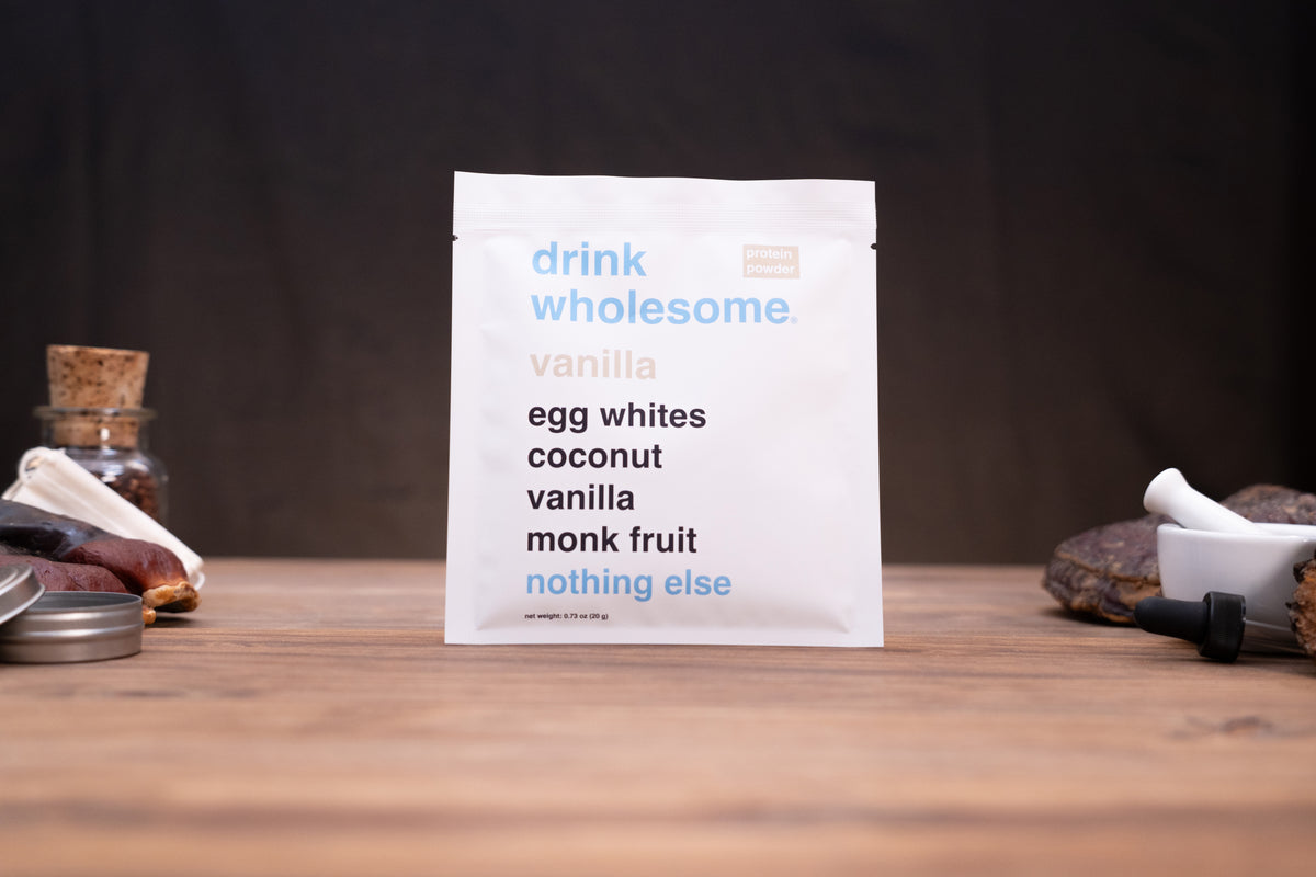 A white pouch of 'drink wholesome vanilla' drink mix stands on a wooden table against a dark background, styled with small ingredients like a cork stopper, dried fruit, and a mortar and pestle.