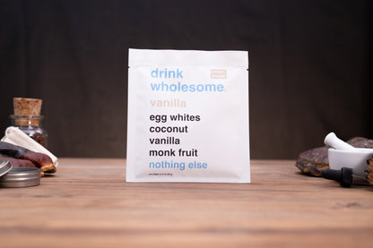 A white pouch of 'drink wholesome vanilla' drink mix stands on a wooden table against a dark background, styled with small ingredients like a cork stopper, dried fruit, and a mortar and pestle.