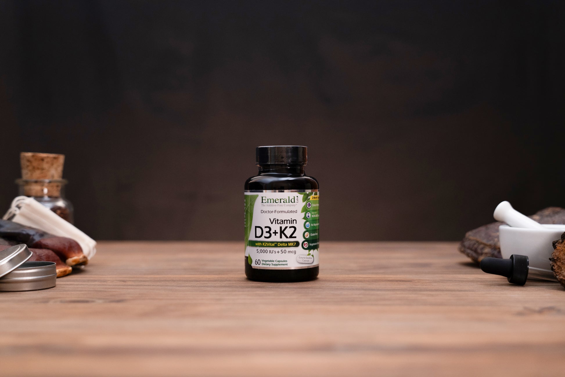 A bottle of Emerald Vitamin D3+K2 dietary supplement standing on a wooden tabletop, with apothecary-style props blurred in the background.