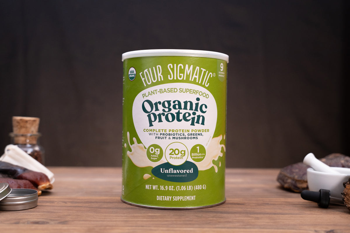 A canister of Four Sigmatic Organic Protein powder sits on a wooden surface with a dark, moody background. The canister is light green with white and dark green text, and is flanked by small, blurred rustic props on both sides.