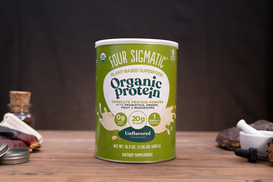 A canister of Four Sigmatic Organic Protein powder sits on a wooden surface with a dark, moody background. The canister is light green with white and dark green text, and is flanked by small, blurred rustic props on both sides.