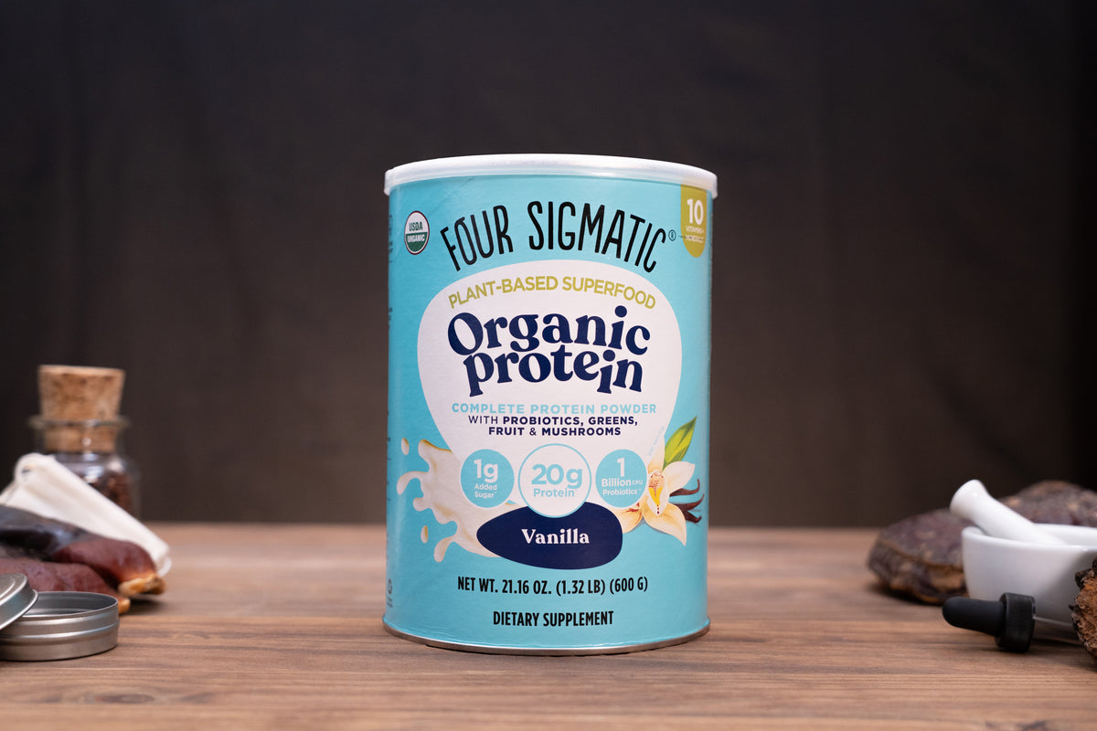 A canister of Four Sigmatic Organic Protein powder in Vanilla flavor, displayed on a wooden surface with rustic props like a mortar and pestle, dried herbs, and a small bottle in the background.