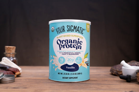 A canister of Four Sigmatic Organic Protein powder in Vanilla flavor, displayed on a wooden surface with rustic props like a mortar and pestle, dried herbs, and a small bottle in the background.