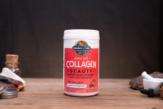 A white plastic tub of Garden of Life Collagen Beauty powder with a red and white label, set on a wooden table with herbal props in the background.