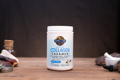 A white plastic tub of Garden of Life Collagen Creamer powder stands on a wooden surface against a dark backdrop. The label reads 'Garden of Life' and 'COLLAGEN CREAMER' with 'Flavored with Coconut Milk' and 'Peanut, Banana, and Coconut' visible.