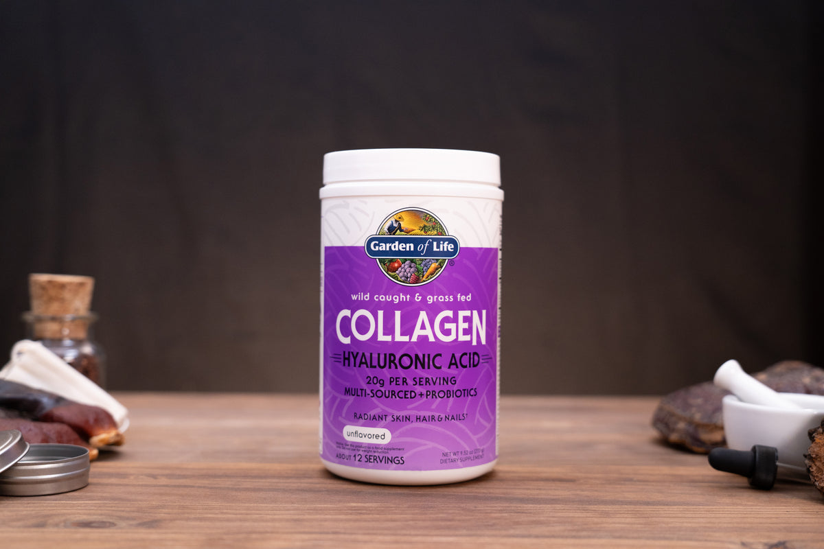 A white plastic bottle of Garden of Life Collagen + Hyaluronic Acid supplement, featuring a purple label, set on a wooden surface with rustic herbal props in the background.