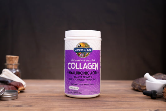 A white plastic bottle of Garden of Life Collagen + Hyaluronic Acid supplement, featuring a purple label, set on a wooden surface with rustic herbal props in the background.