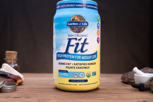 A canister of Garden of Life Fit Raw Organic High Protein powder, vanilla flavor, displayed on a wooden surface with herbal and supplement props in the background.