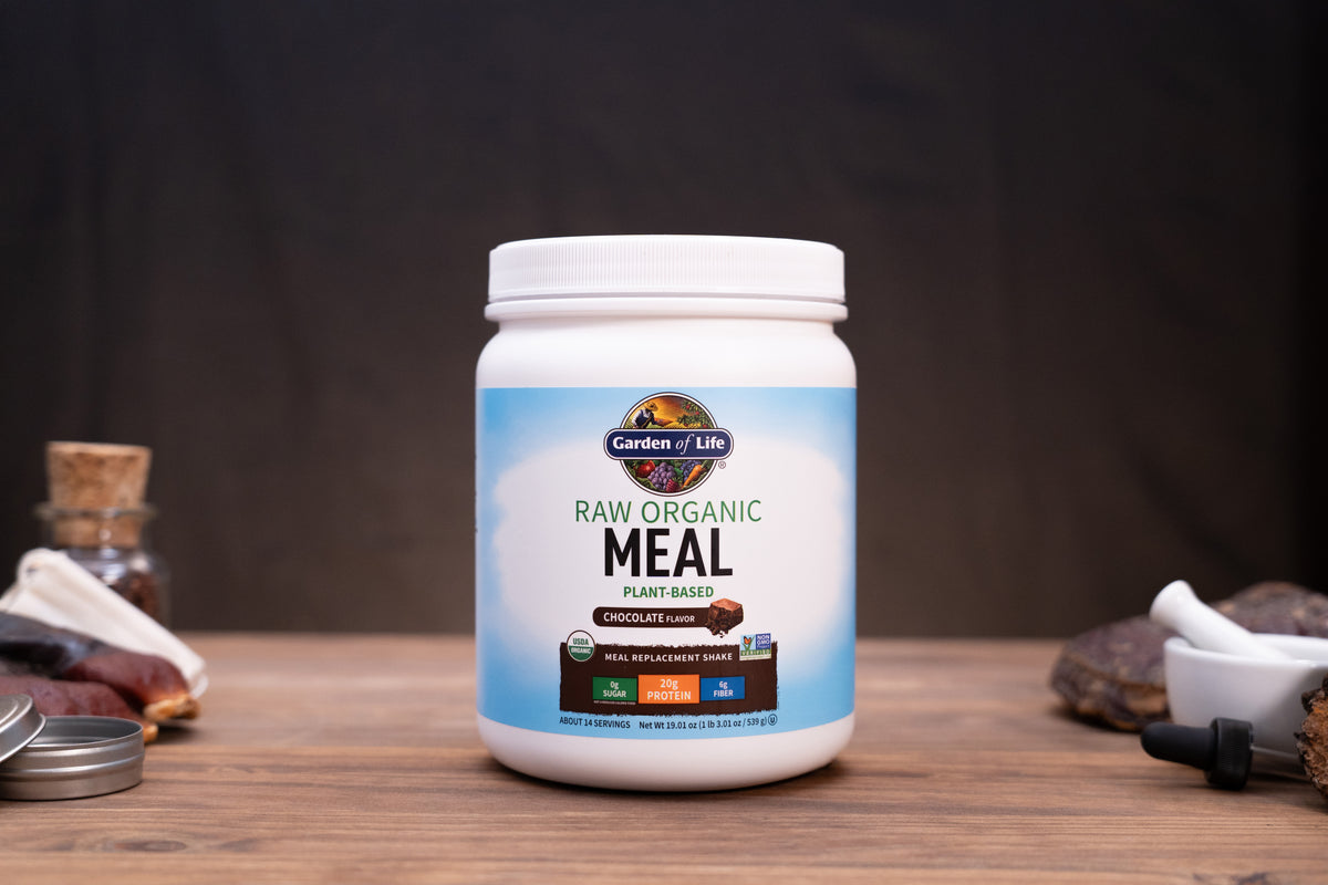 A white plastic jar of Garden of Life Raw Organic Meal powder, set on a rustic wooden table with dark background and small natural props.