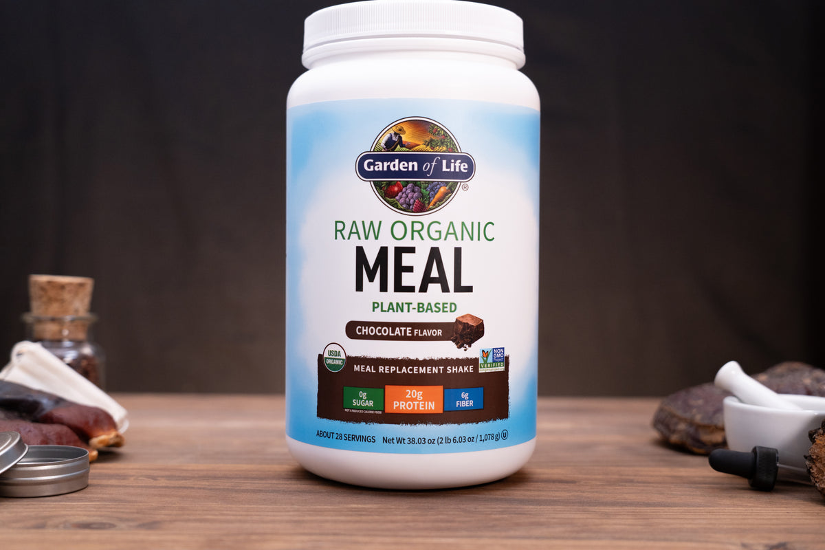A white tub of Garden of Life Raw Organic Meal plant-based chocolate flavor meal replacement shake, displayed on a wooden surface with rustic ingredients in the background.