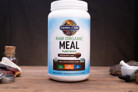 A white tub of Garden of Life Raw Organic Meal plant-based chocolate flavor meal replacement shake, displayed on a wooden surface with rustic ingredients in the background.