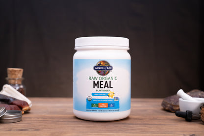 A white plastic tub of Garrow's Labs Raw Organic Meal Plant Based supplement, displayed on a rustic wooden table with dark background and small natural props.