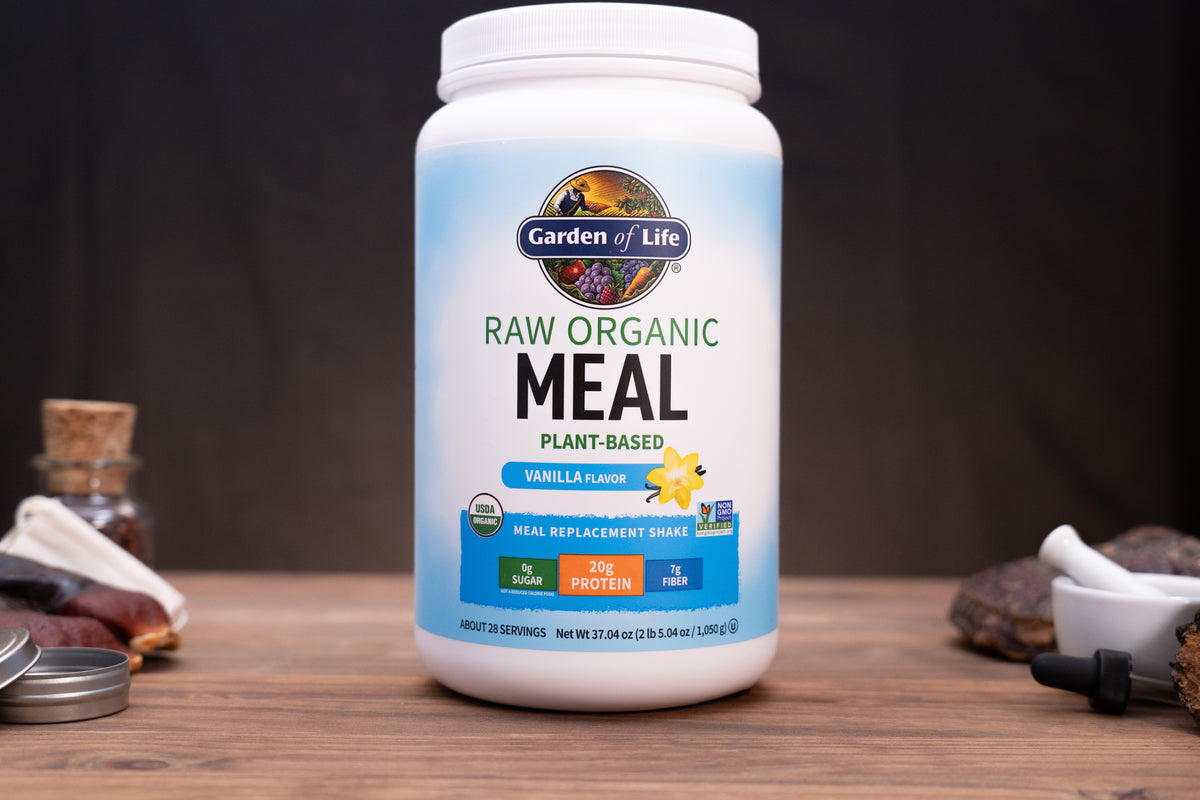 A white tub of Garden of Life Raw Organic Meal Plant-Based Vanilla Flavour meal replacement shake powder, displayed on a wooden surface with blurred natural ingredients in the background.