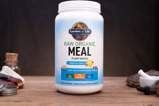 A white tub of Garden of Life Raw Organic Meal Plant-Based Vanilla Flavour meal replacement shake powder, displayed on a wooden surface with blurred natural ingredients in the background.