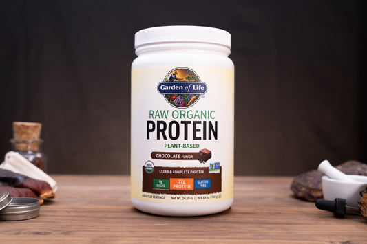 A white tub of Garden of Life Raw Organic Protein powder in Chocolate Flavors, displayed on a wooden surface with rustic ingredients in the background.