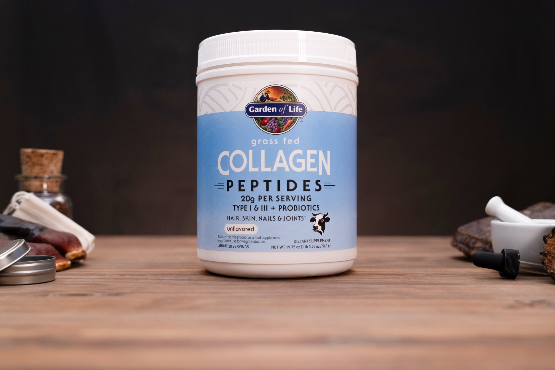 A white plastic tub of Garden of Life Grass Fed Collagen Peptides on a wooden tabletop, surrounded by blurred natural ingredients and a mortar and pestle in the background.