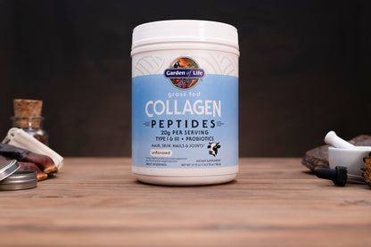 A white plastic tub of Garden of Life Grass Fed Collagen Peptides on a wooden tabletop, surrounded by blurred natural ingredients and a mortar and pestle in the background.