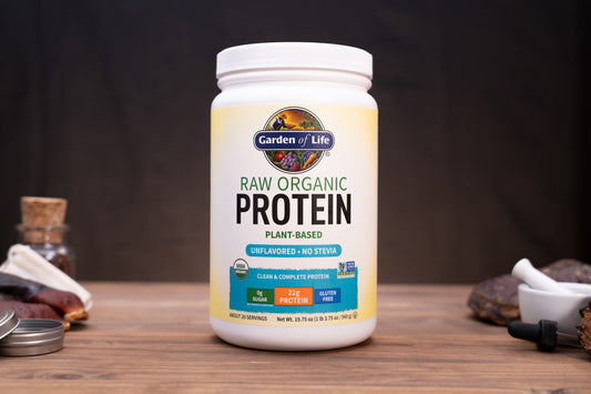 A white tub of Garden of Life Raw Organic Protein powder stands on a wooden surface against a dark background, with small kitchen or supplement props visible around it.