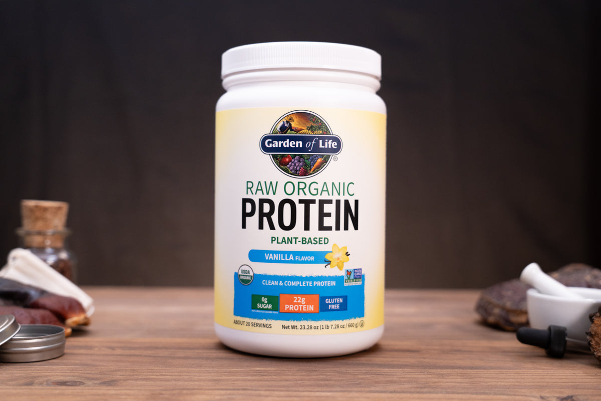 A white tub of Garden of Life Raw Organic Protein Vanilla flavor, displayed on a wooden surface with a dark, blurred background and small kitchen props on either side.