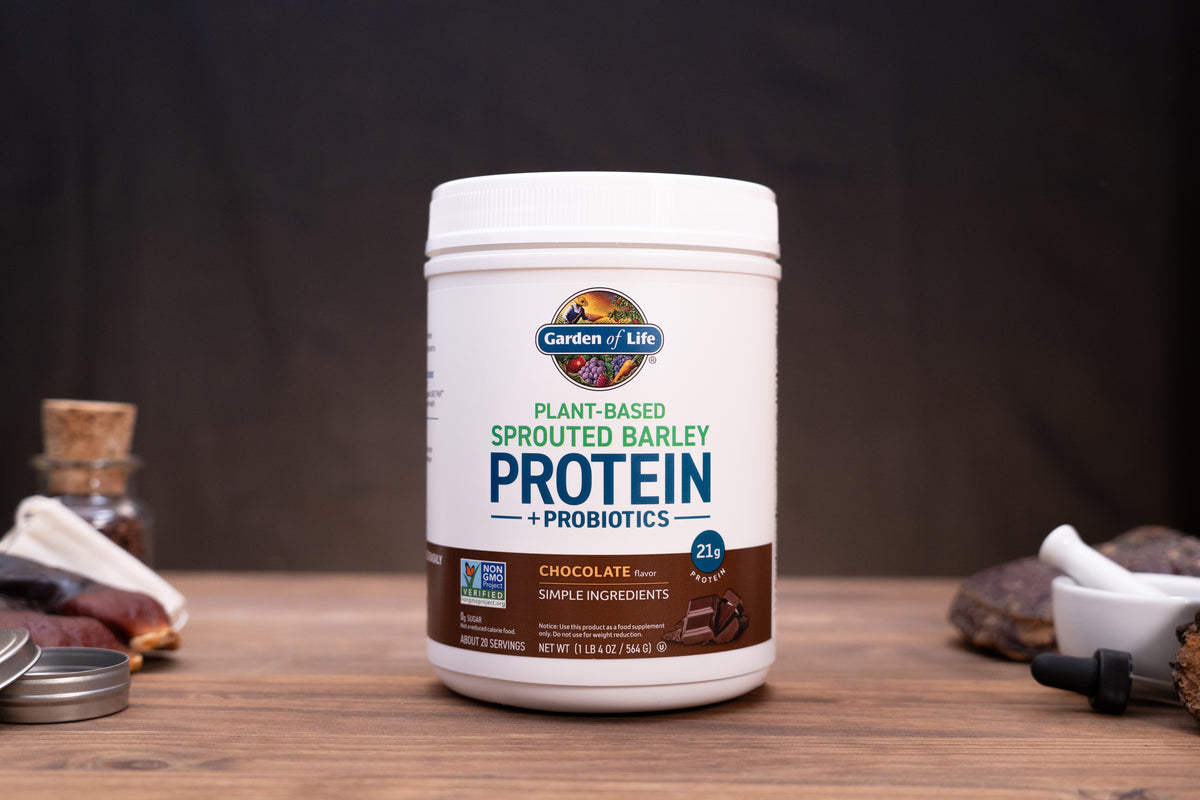 A white tub of Garden of Life Plant-Based Sprouted Barley Protein + Probiotics in Chocolate flavor, displayed on a wooden surface with rustic ingredients in the background.