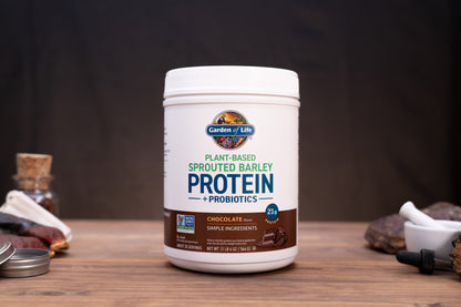 A white tub of Garden of Life Plant-Based Sprouted Barley Protein + Probiotics in Chocolate flavor, displayed on a wooden surface with rustic ingredients in the background.