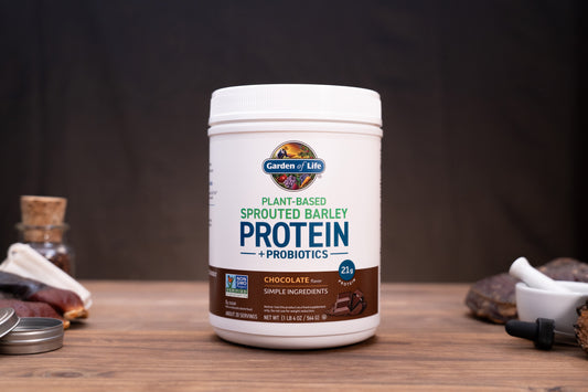 A white tub of Garden of Life Plant-Based Sprouted Barley Protein + Probiotics in Chocolate flavor, displayed on a wooden surface with rustic ingredients in the background.