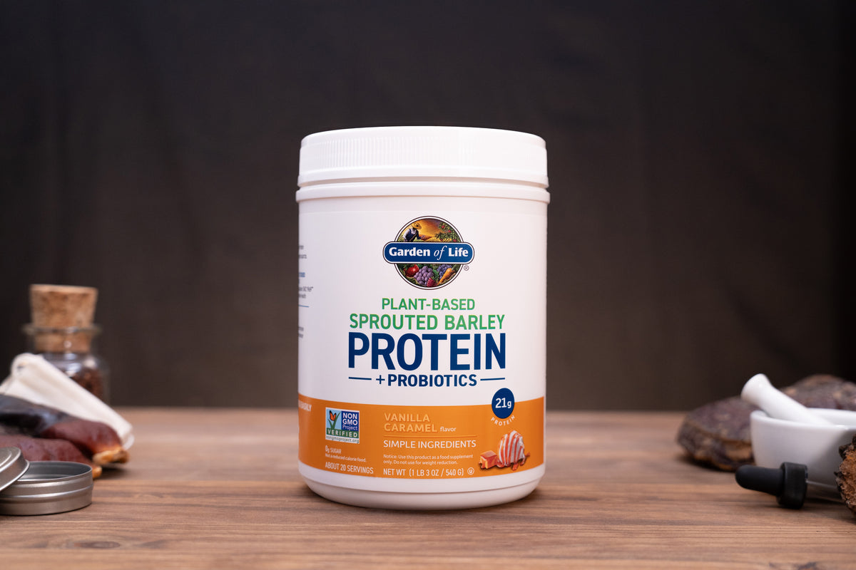 A white tub of Gardens of Life Plant-Based Sprouted Barley Protein + Probiotics in vanilla flavor, displayed on a wooden surface with small natural props in the background.