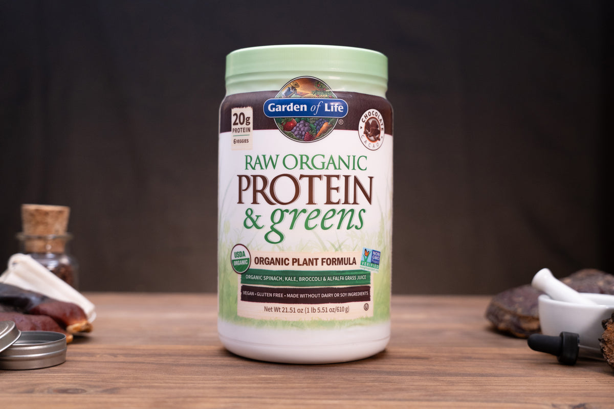 A tub of Garden of Life Raw Organic Protein & Greens powder sits on a wooden surface against a dark background, with small kitchen or supplement props blurred around it.