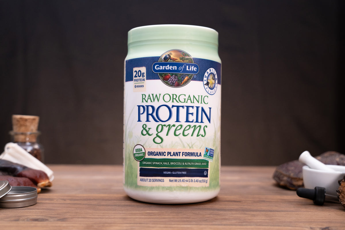 A tub of Garden of Life Raw Organic Protein & Greens powder supplement, displayed on a wooden surface with rustic ingredients in the background.