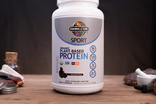 A white tub of Garden of Life Sport Organic Plant-Based Protein powder on a wooden table, surrounded by small kitchen and supplement props against a dark background.