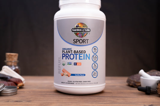 A white plastic tub of Garden of Life Sport Organic Plant-Based Protein powder on a wooden table, with its lid placed beside it. The background is a dark, moody setting with blurred natural elements.