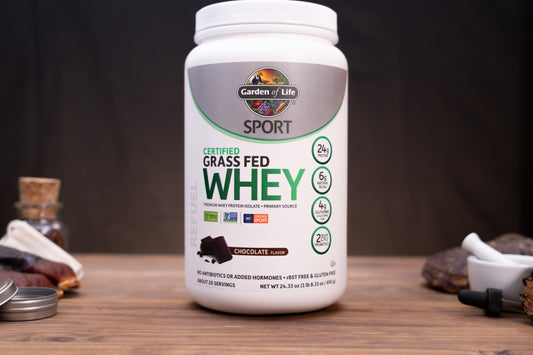 A white tub of Garden of Life Sport Certified Grass Fed Whey in Chocolate Flavor, displayed on a wooden surface with small kitchen props in the background.