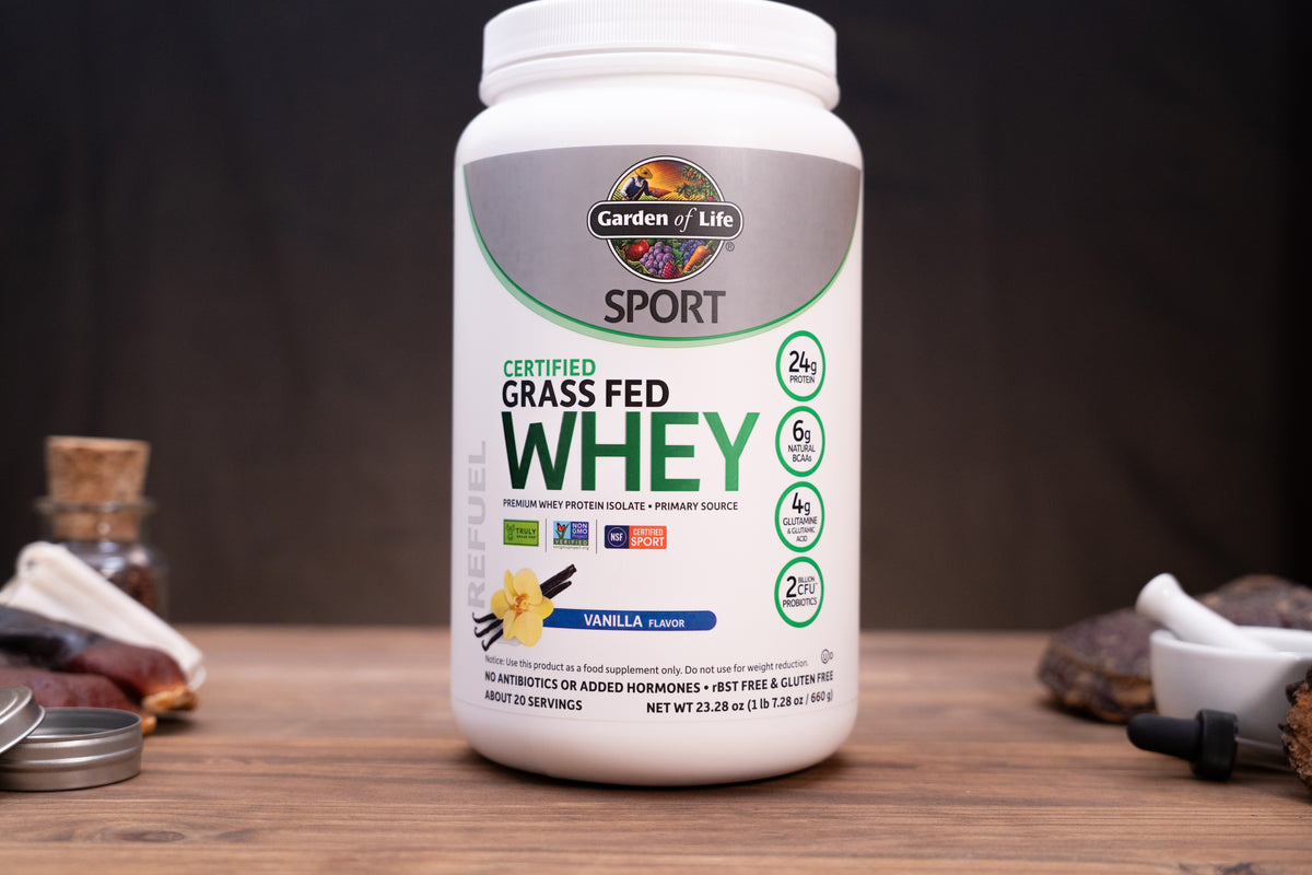 A white tub of Garden of Life Sport Certified Grass Fed Whey in Vanilla Flavor, displayed on a wooden surface with small kitchen props in the background.
