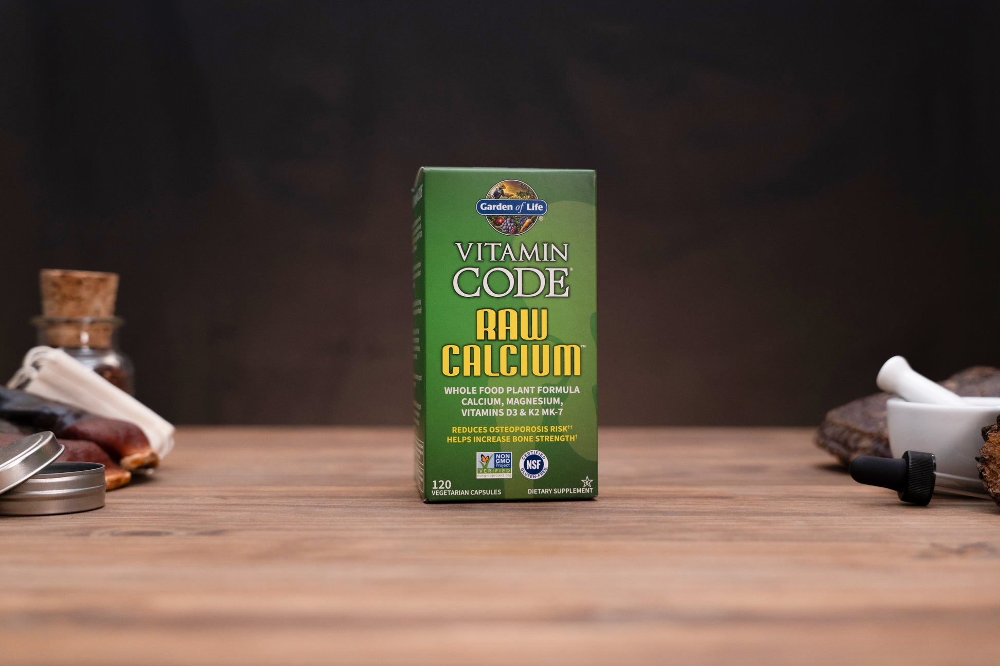 A green box of Garden of Life Vitamin Code Raw Calcium stands on a wooden surface against a dark, blurred background, with small tabletop props visible on both sides.