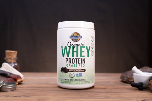 A white tub of Garden of Life Organic Whey Protein Grass Fed Chocolate Cocoa flavor, displayed on a wooden surface with rustic props in the background.