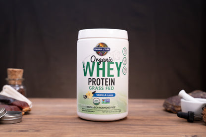 A white tub of Garden of Life Organic Whey Protein Grass Fed Vanilla Flavour, displayed on a wooden surface with small rustic props in the background.