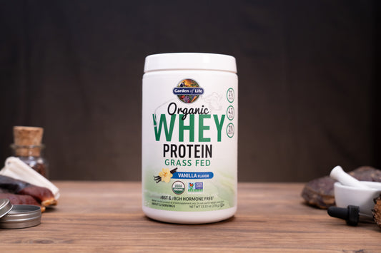 A white tub of Garden of Life Organic Whey Protein Grass Fed Vanilla Flavour, displayed on a wooden surface with small rustic props in the background.