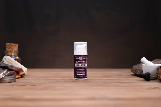 A small bottle of Gads & Real Ice Lotion stands centered on a wooden tabletop against a dark brown background, with blurred herbal and cosmetic props visible on both sides.