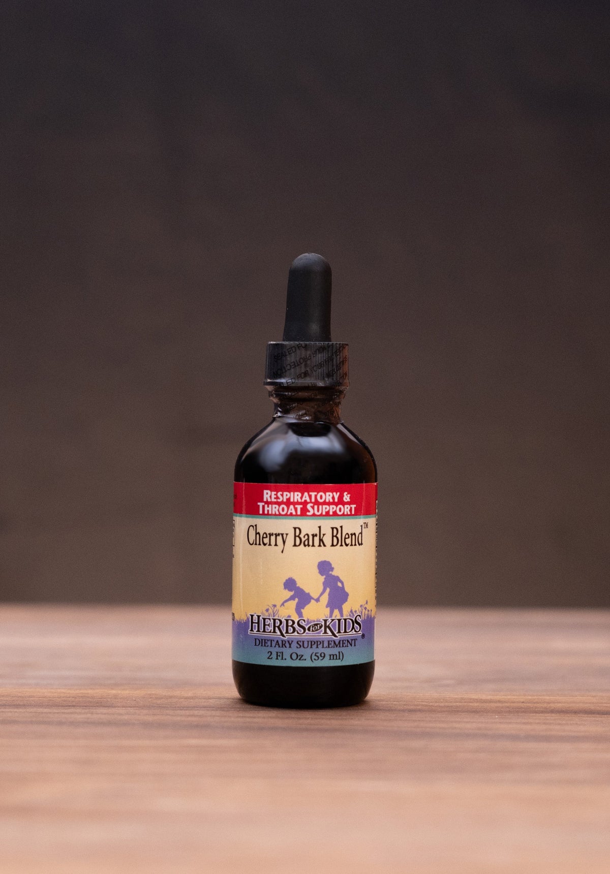 A small amber glass dropper bottle labeled Cherry Bark Blend stands centered on a wooden tabletop against a dark brown backdrop. The bottle is surrounded by rustic props, including a cork stopper, a metal tin lid, a white mortar and pestle, and a small jar, creating a styled product presentation.